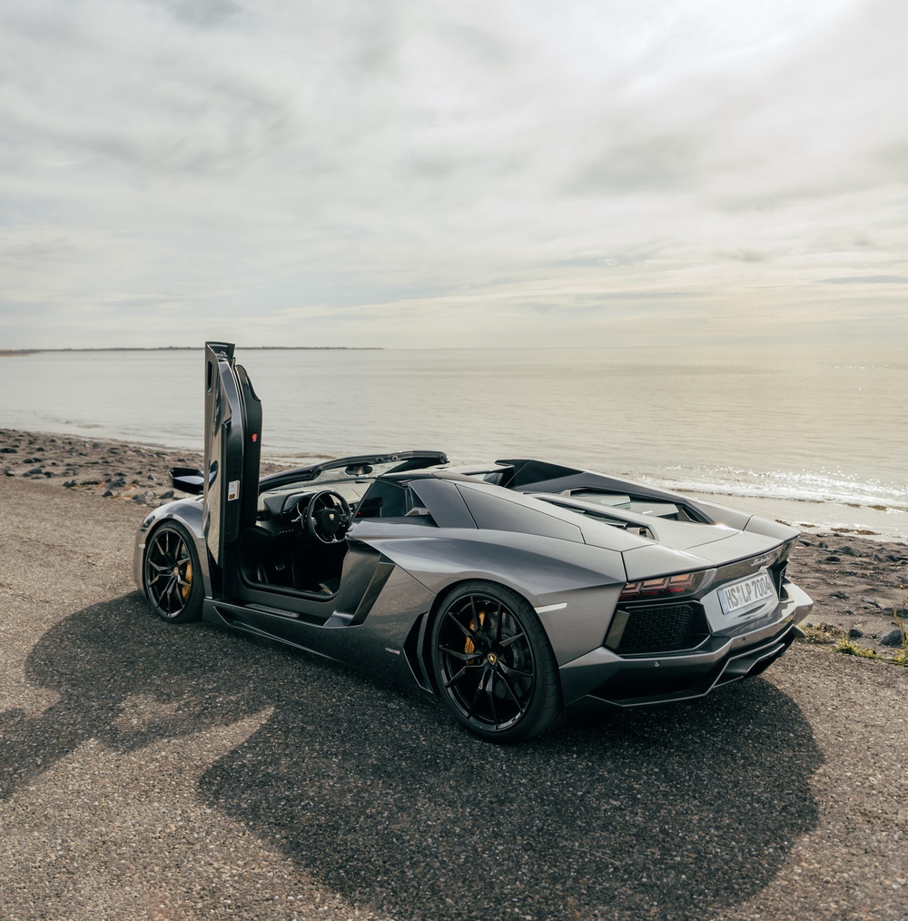Lamborghini Aventador Roadster in de kleur grijs met de deur open op een kade langs het strand.